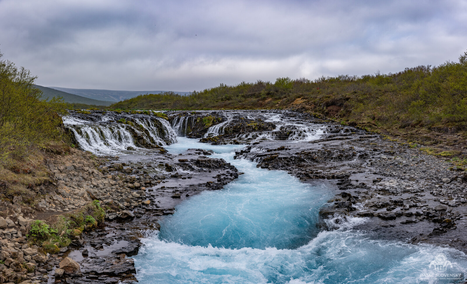 Panorama of Brúarfoss, electric-blue glacial water spilling over dark lava ledges
