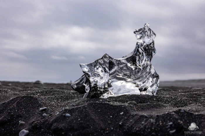 Sculpted clear ice fragment glowing on black volcanic sand, low angle