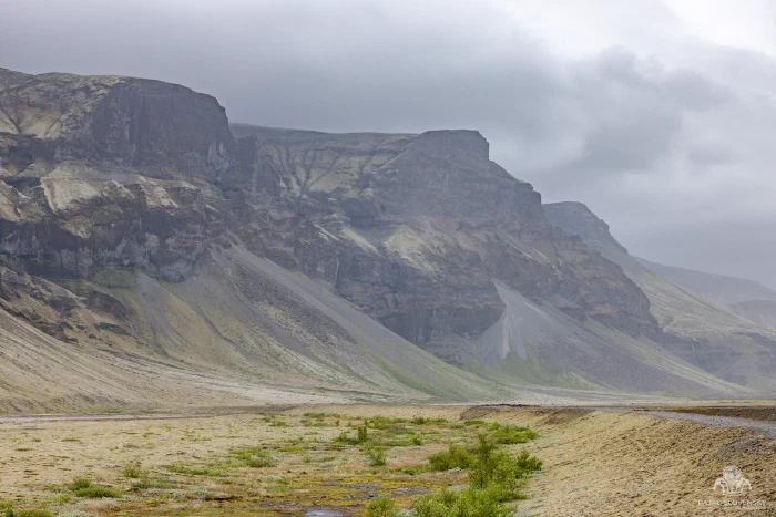 Rain-hazed layered cliffs and scree fans above sandy valley floor