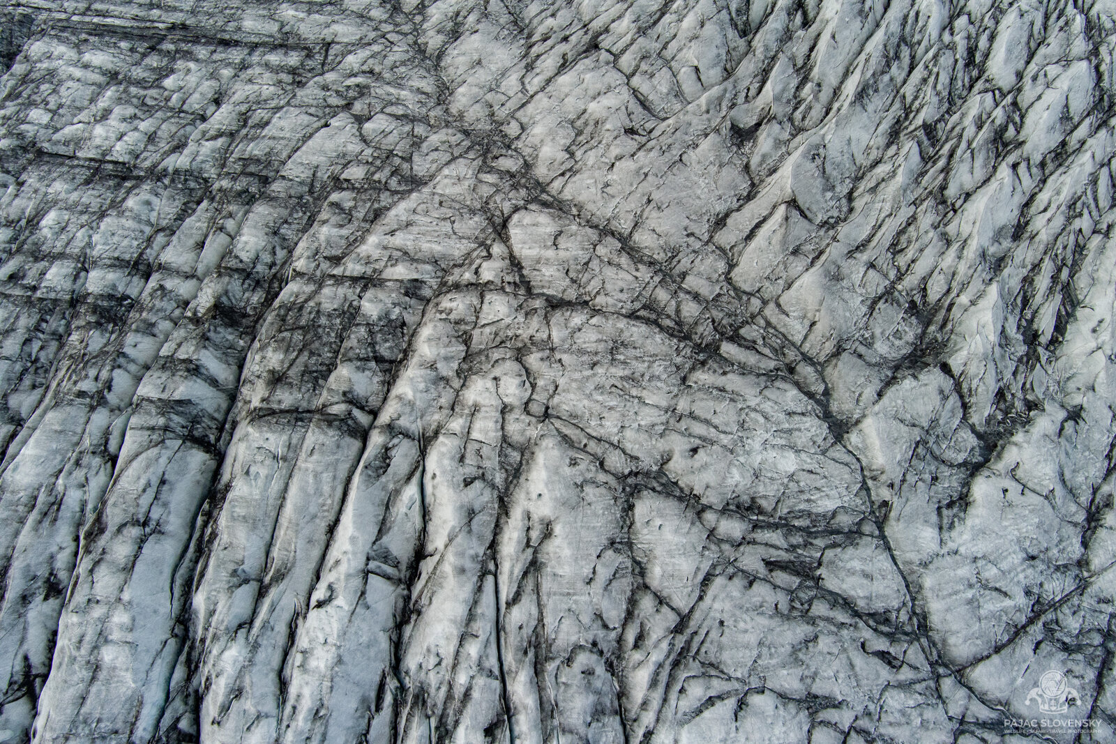 Aerial of a white glacier crisscrossed by dark crevasses and streaks of volcanic ash