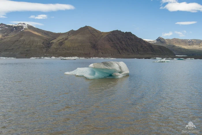 Turquoise iceberg drifting on silty grey glacier lagoon beneath brown mountains