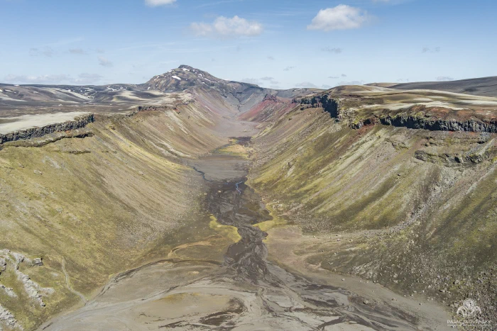 Drone view down moss-green canyon, dark braided stream threading valley floor