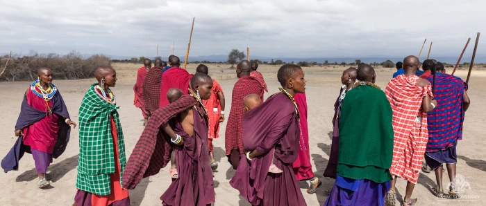 Maasai women in layered shukas and beaded collars, one carrying infant