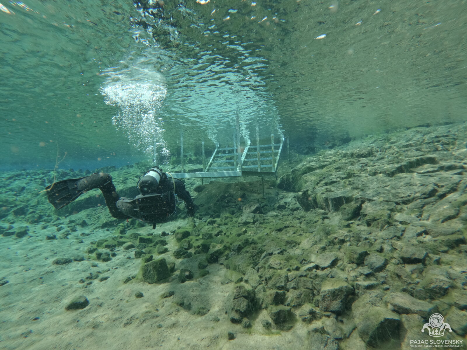 Diver approaching the metal exit stairs of Silfra through shallow water, trailing bubbles
