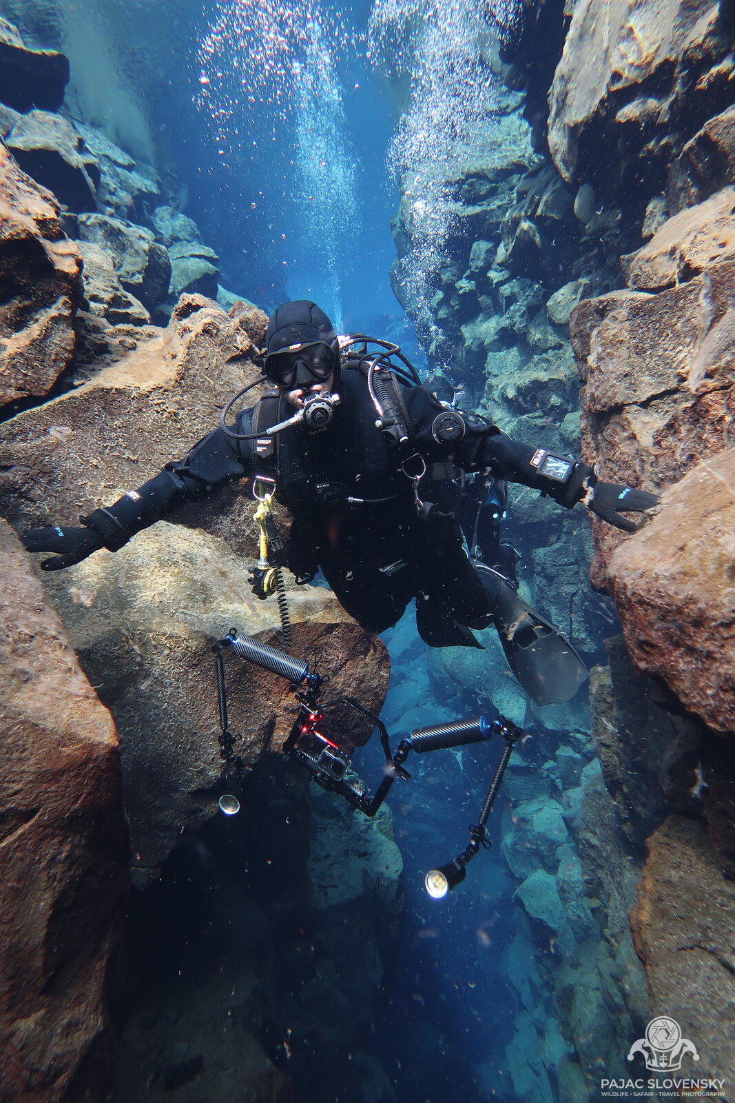 Drysuit diver in the narrow Silfra fissure with arms outstretched, touching both rock walls at once
